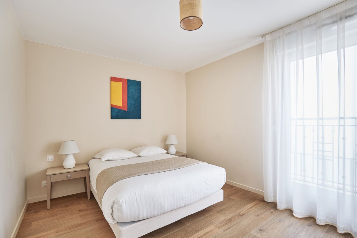 Ein Schlafzimmer mit einem Bett, zwei Nachttischen, einer Lampe auf jedem Tisch, einem Wandbild und einem Fenster mit transparenten Vorhängen.