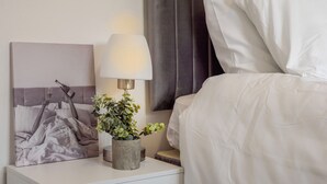 A neatly made bed with white linens, a bedside lamp, a framed artwork, and a small potted plant on a white nightstand. - Cosy 2-bedroom house with Parking (Salford)