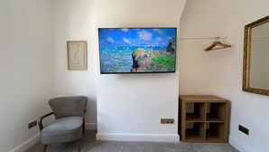 Una habitación con un sillón gris, un cuadro colgado con una escena costera, una estantería de madera y un espejo en la pared.