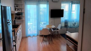 Een kleine keuken met een koelkast, magnetron en witte kastjes. Er is een eethoek met een tafel en stoelen, en een aan de muur bevestigde televisie.