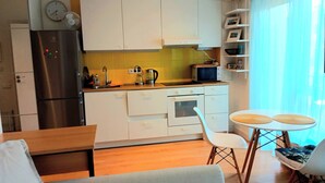 Een kleine keuken met een koelkast, magnetron en fornuis. Er is een eethoek met een tafel en stoelen.