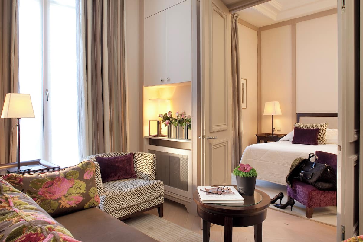 A hotel room with a bed, a sofa, a chair, a small table with a book and a plant, and a nightstand with a lamp.