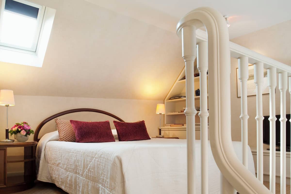 A bedroom with a bed, two pillows, a nightstand with a lamp, a window, and a staircase with white balusters.
