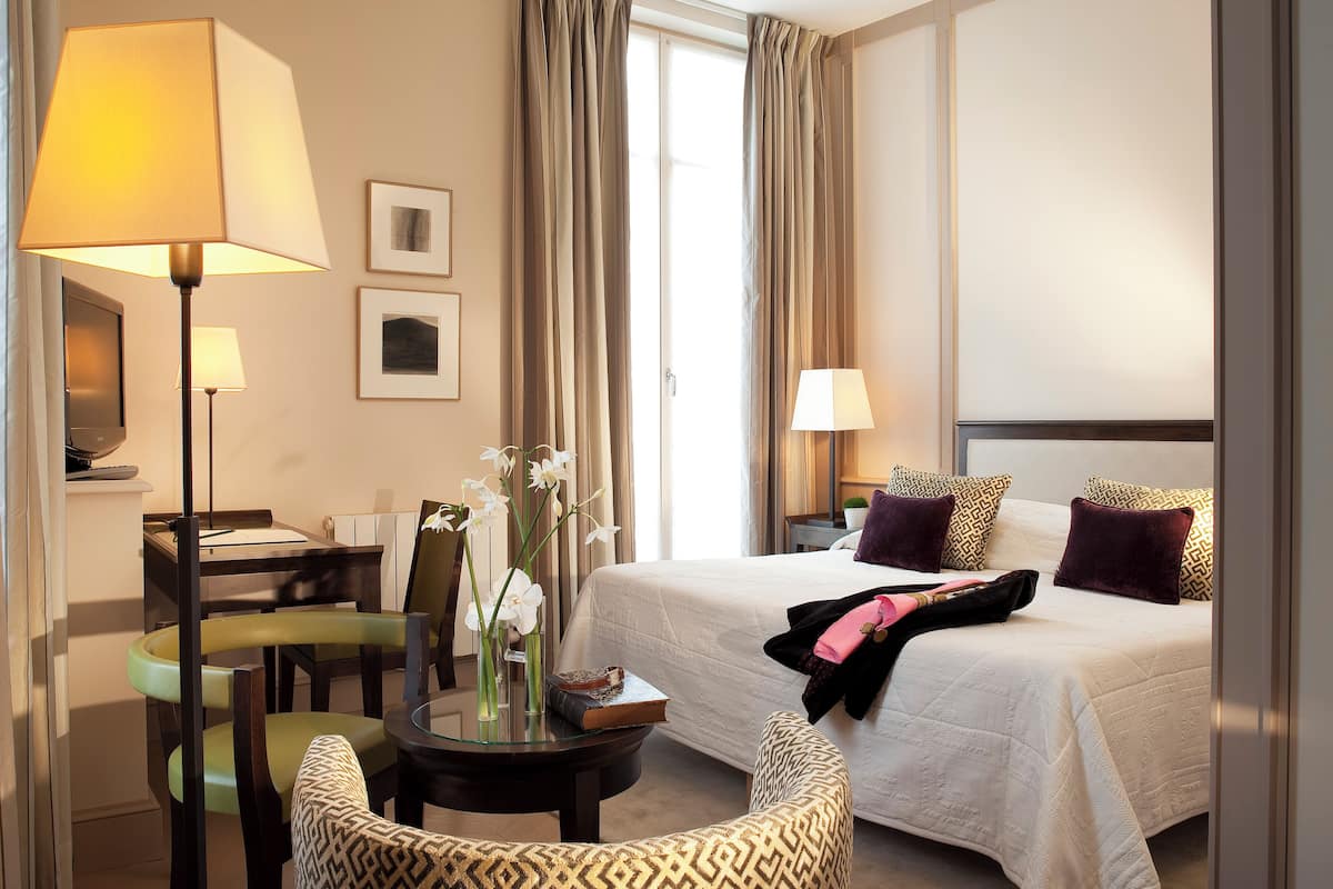A hotel room with a bed, two bedside lamps, a desk with a chair, a small table with a vase of flowers, and a window with curtains.