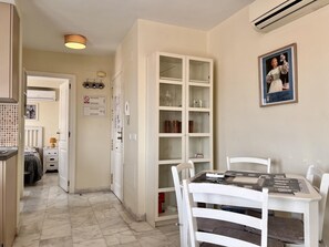 Um corredor estreito com área de jantar, destacando uma mesa e cadeiras brancas, um armário com portas de vidro e um ar-condicionado fixado na parede.