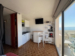Habitación compacta con kitchenette, un pequeño comedor, televisor montado en la pared y vista a la playa a través de amplias ventanas.