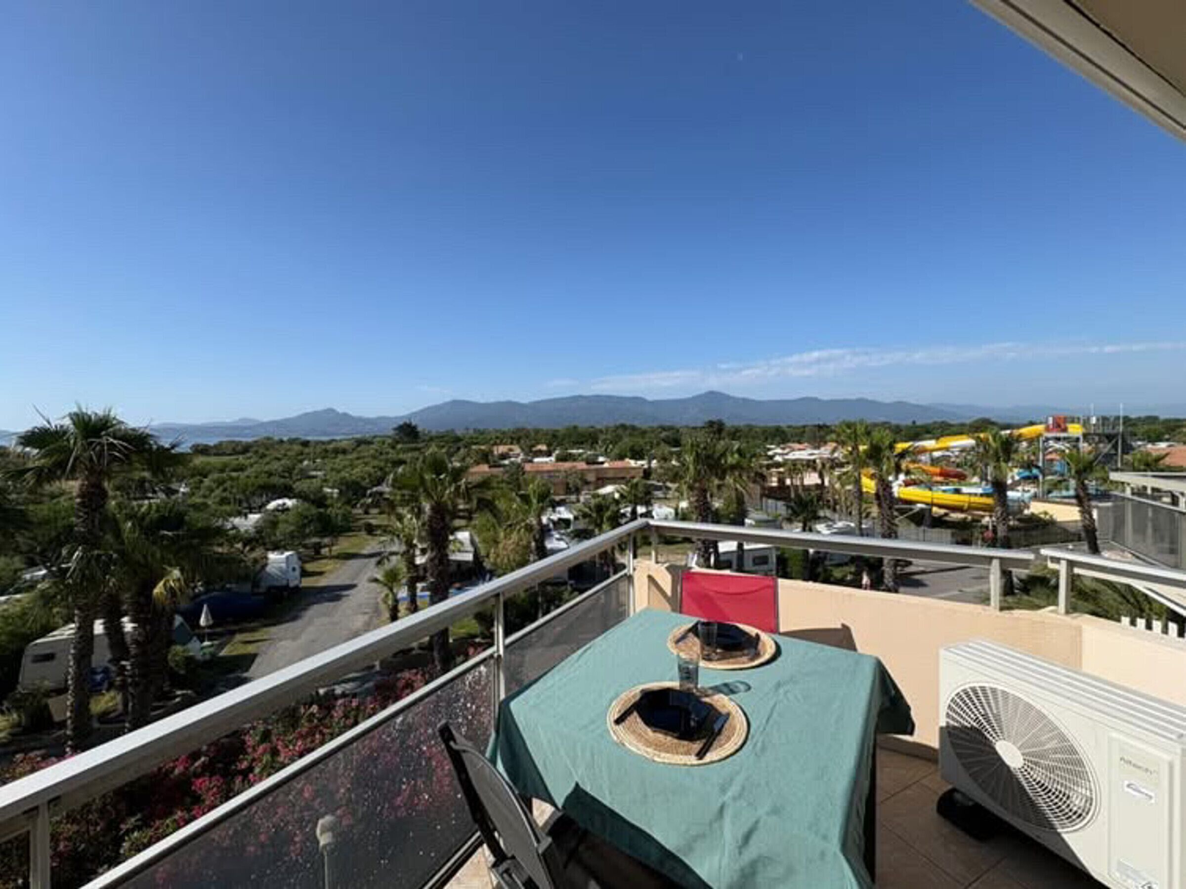 Ein Balkon mit Tisch, einem Ventilator und Blick auf einen Wasserpark und Berge.