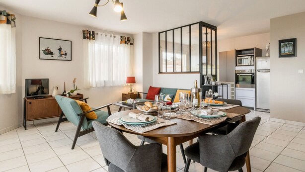A modern dining area with a wooden table, grey chairs, and a kitchen in the background.