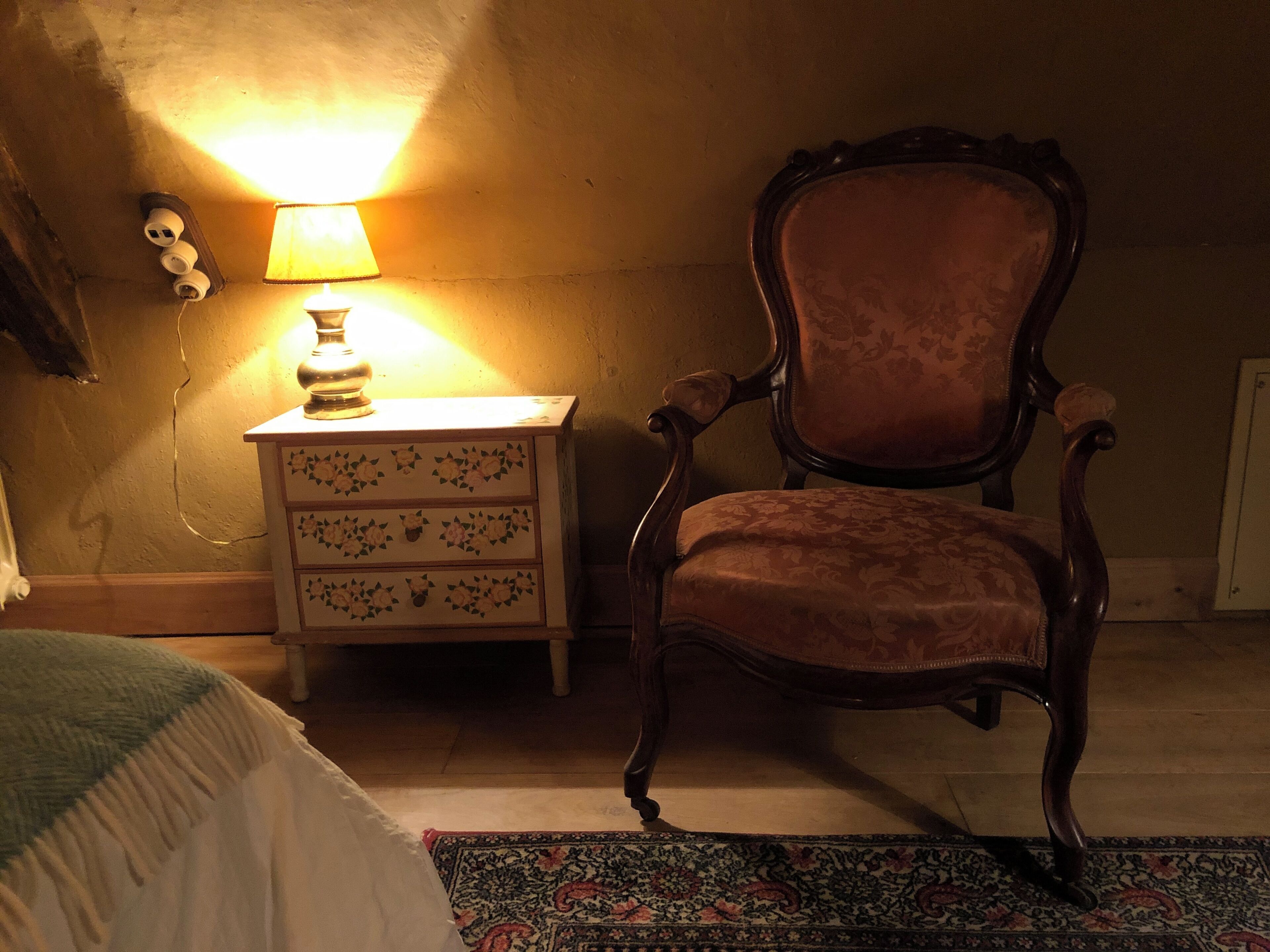 A room with a wooden chair, a bedside table with a lamp, and a patterned rug.