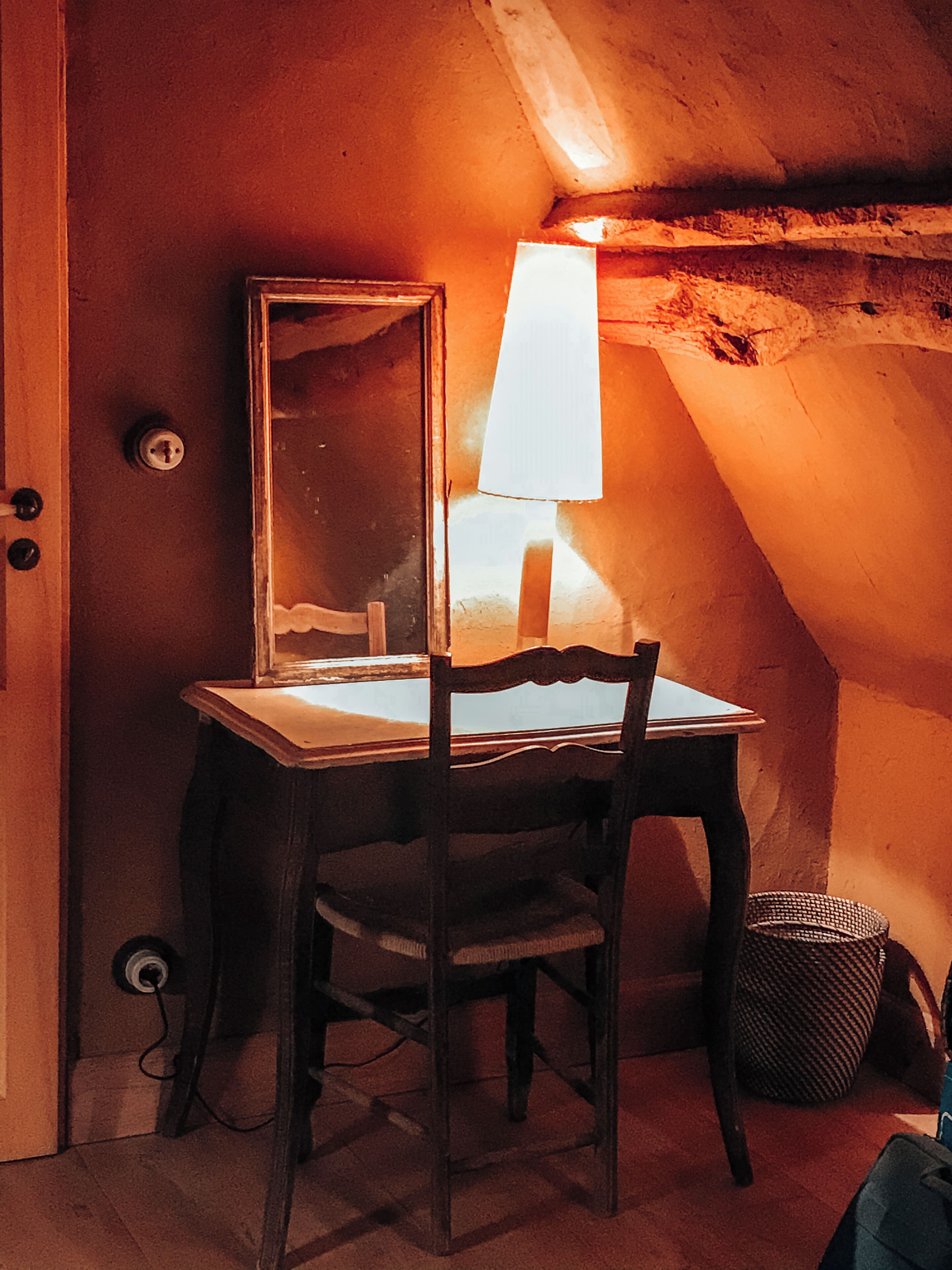 Una stanza piccola, scarsamente illuminata, con una scrivania in legno, una sedia, una lampada e uno specchio.