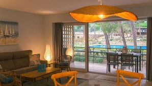 Un salon comprenant un canapé, une table basse et des fauteuils, un coin repas avec une table et des chaises, et une grande fenêtre donnant sur une piscine et des arbres.
