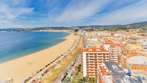 A coastal cityscape with a wide beach, a row of parked cars, and a prominent multi-story building with orange roofs.