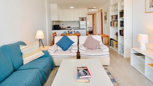 A compact living space with a sofa, a coffee table, a bookshelf, and a kitchenette in the background.