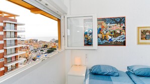 A bedroom with a large window, a bed with blue bedding, and two framed artworks on the wall.