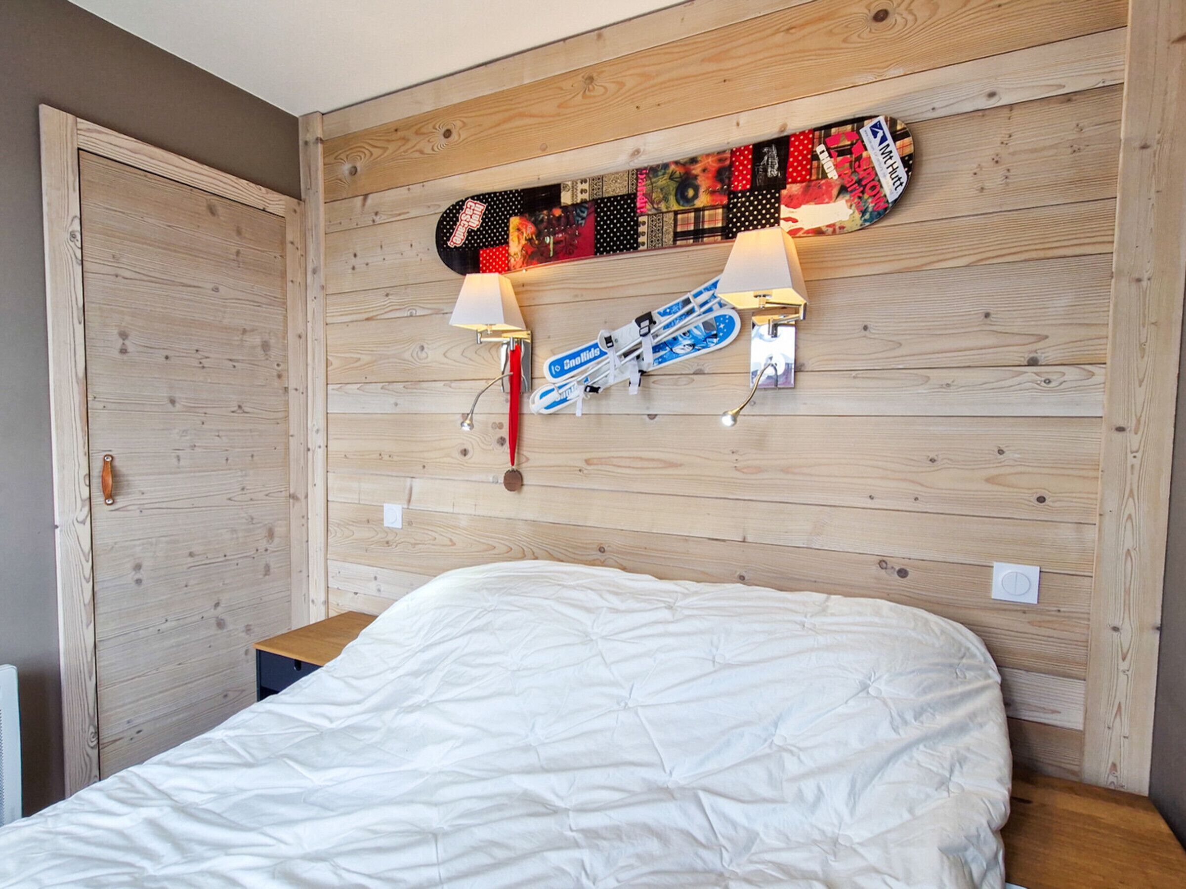 Ein Schlafzimmer mit Holz-Kopfteil, einem Bett mit weißer Bettwäsche, zwei Wandlampen und einem Skateboard mit verschiedenen Abzeichen und Aufklebern.