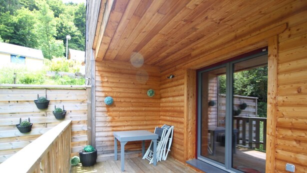 Une terrasse en bois avec une table et des chaises, des plantes en pot et une porte-fenêtre coulissante donnant sur un espace extérieur.