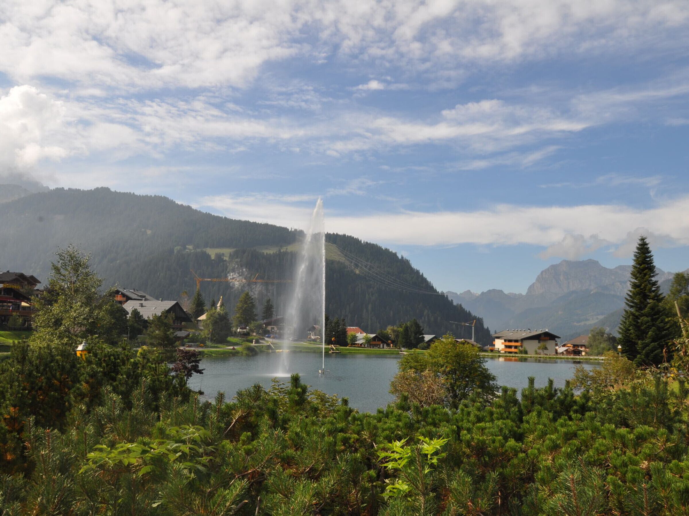 A lakeside view with a fountain, surrounded by houses and mountains.