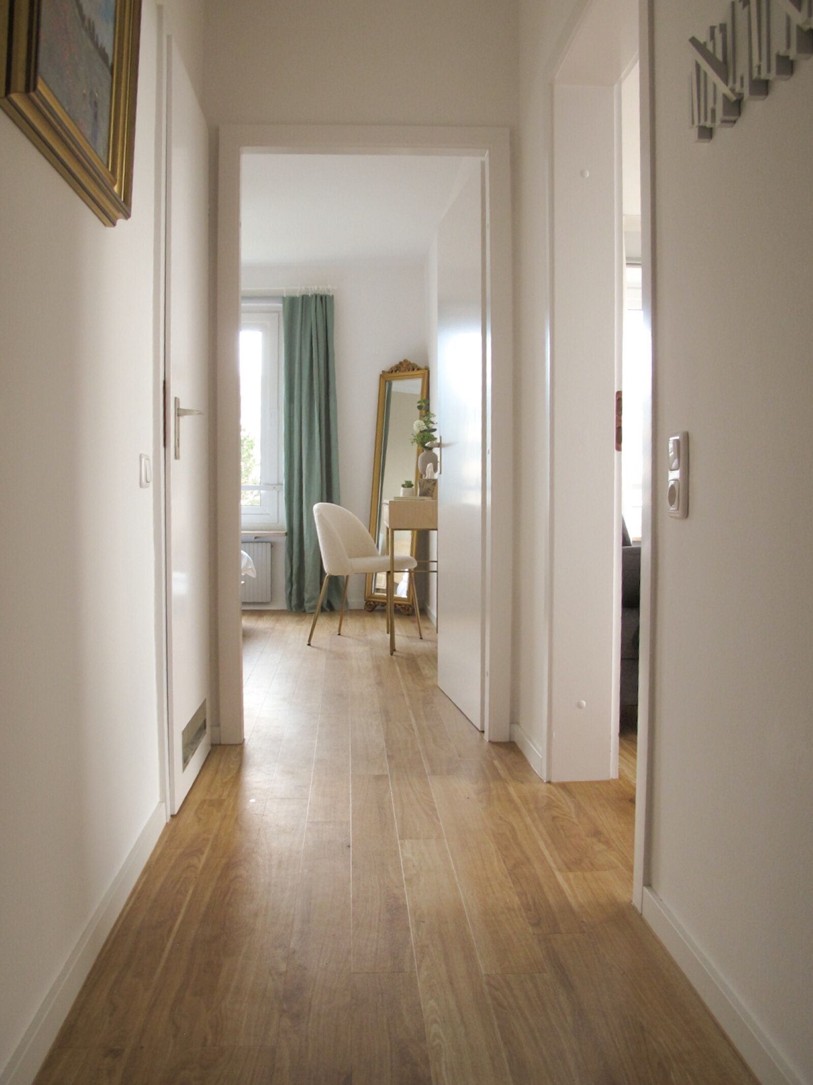 A hallway with wooden flooring leading to a room with a desk, chair, and a mirror.