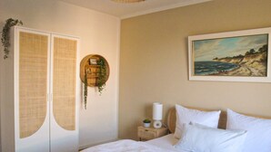 Un dormitorio con cama, una mesita de noche, un estante en la pared con plantas y un cuadro de una escena costera.