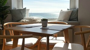 Un comedor con una mesa y sillas, vista al mar a través de una ventana y una planta en maceta.