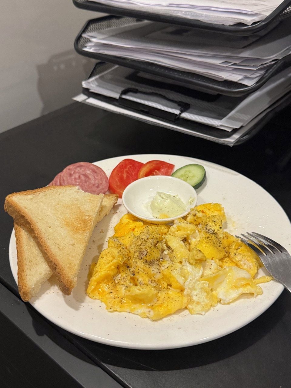 A plate of scrambled eggs, toast, tomatoes, cucumber, and a small bowl of butter.