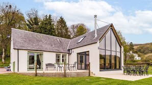 A modern house with a large glass extension, a patio with outdoor furniture, and a well-maintained lawn.