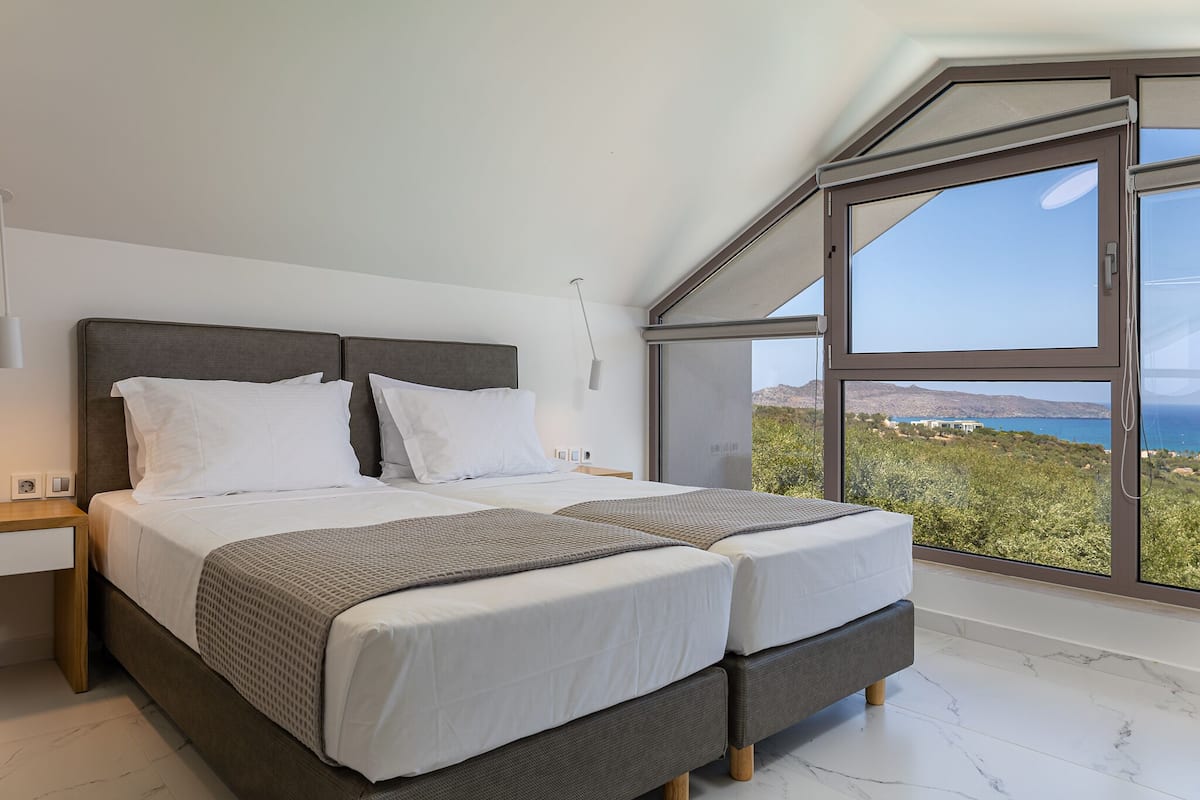 A modern bedroom with two beds, a large window offering a scenic view, and a minimalist design.