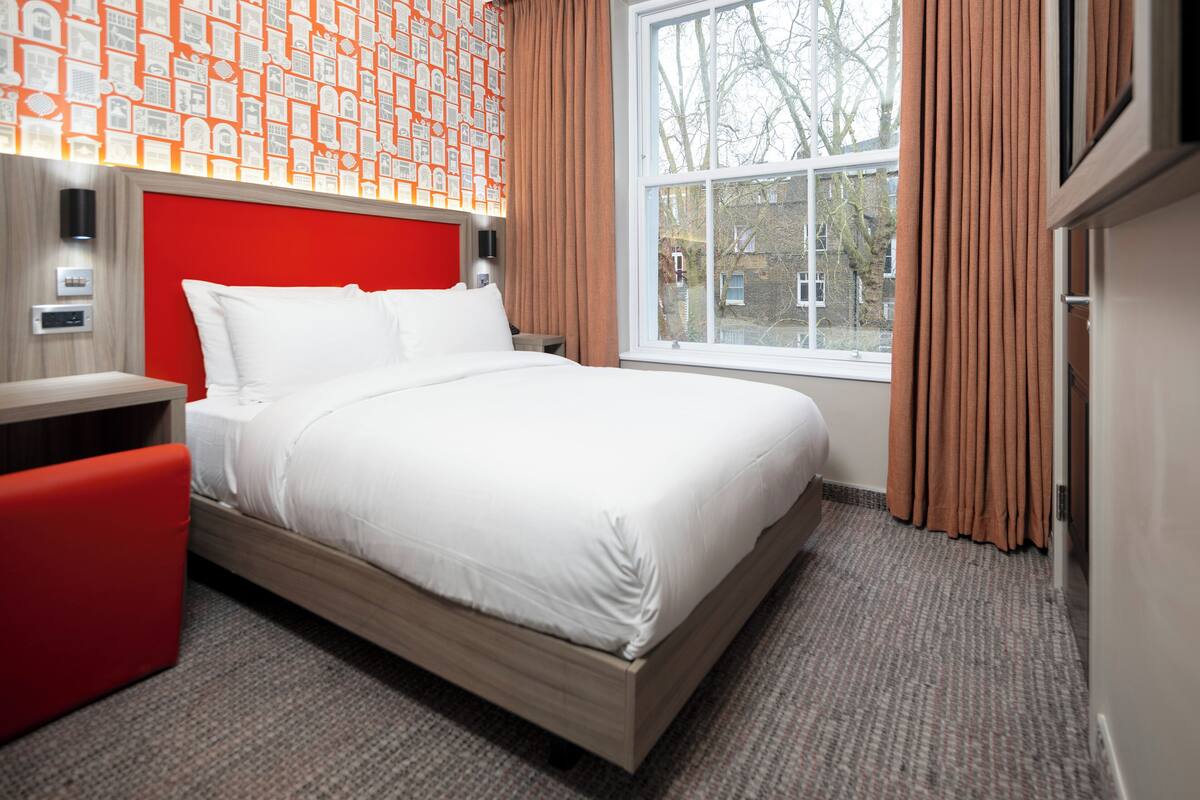 A neatly made bed in a hotel room with a red headboard, a window with curtains, and a patterned wallpaper.
