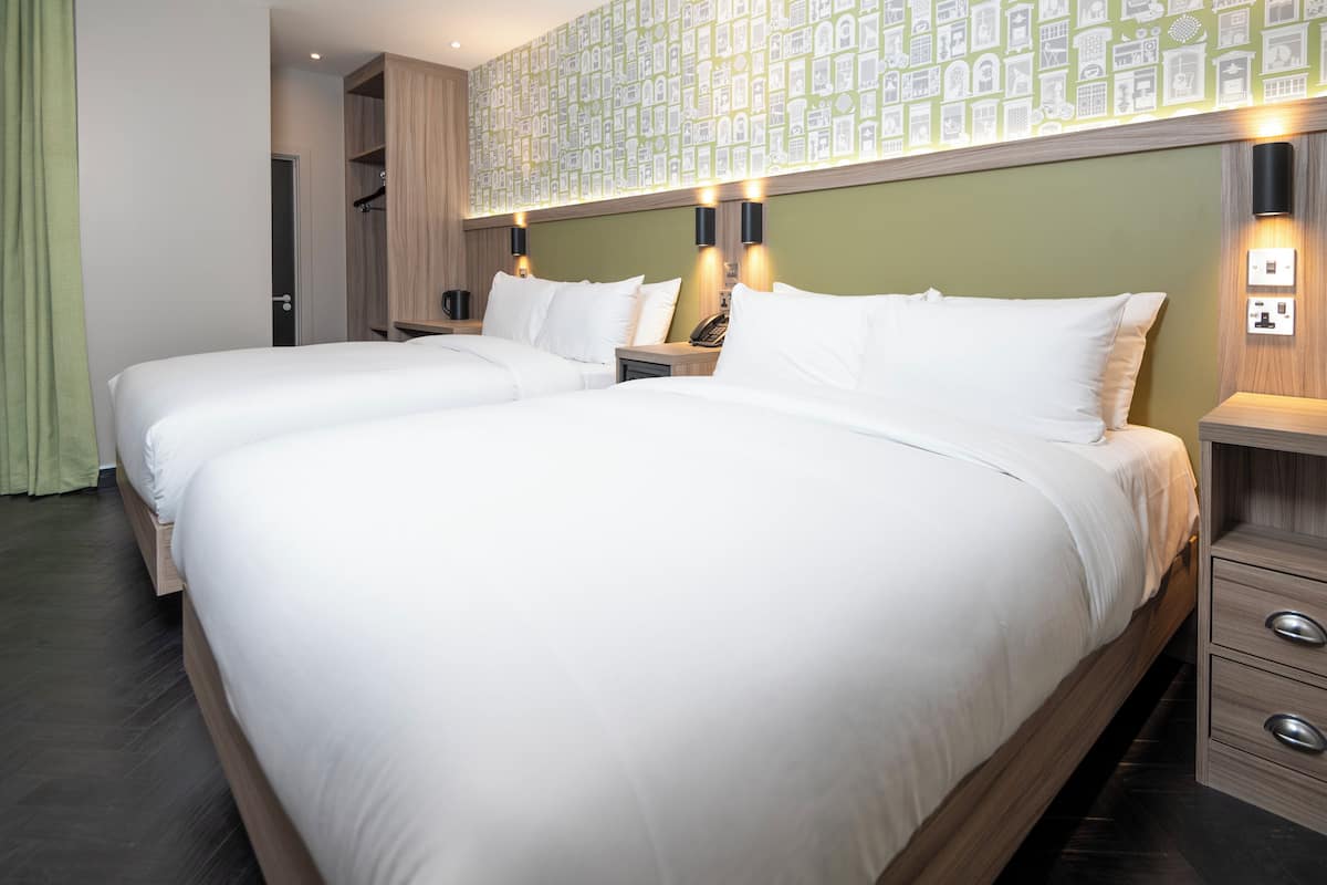 A hotel room with two beds, a wardrobe, a bedside table with a lamp, and a wall-mounted light fixture.