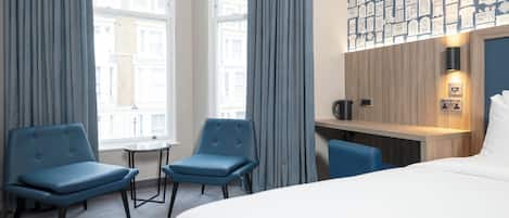 A hotel room with a large bed, two blue armchairs, a small table, a desk with a kettle, and a view of buildings through large windows.