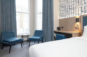 Ein Hotelzimmer mit einem großen Bett, zwei blauen Sessel, einem kleinen Tisch, einem Schreibtisch mit Wasserkocher und Blick auf Gebäude durch große Fenster.