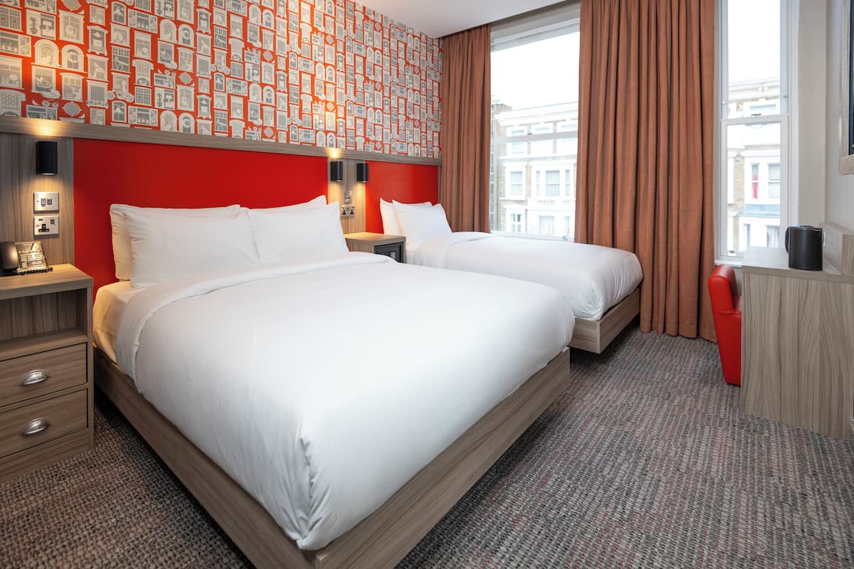 A hotel room with two beds, a red wall with a patterned wallpaper, a large window with brown curtains, and a carpeted floor.