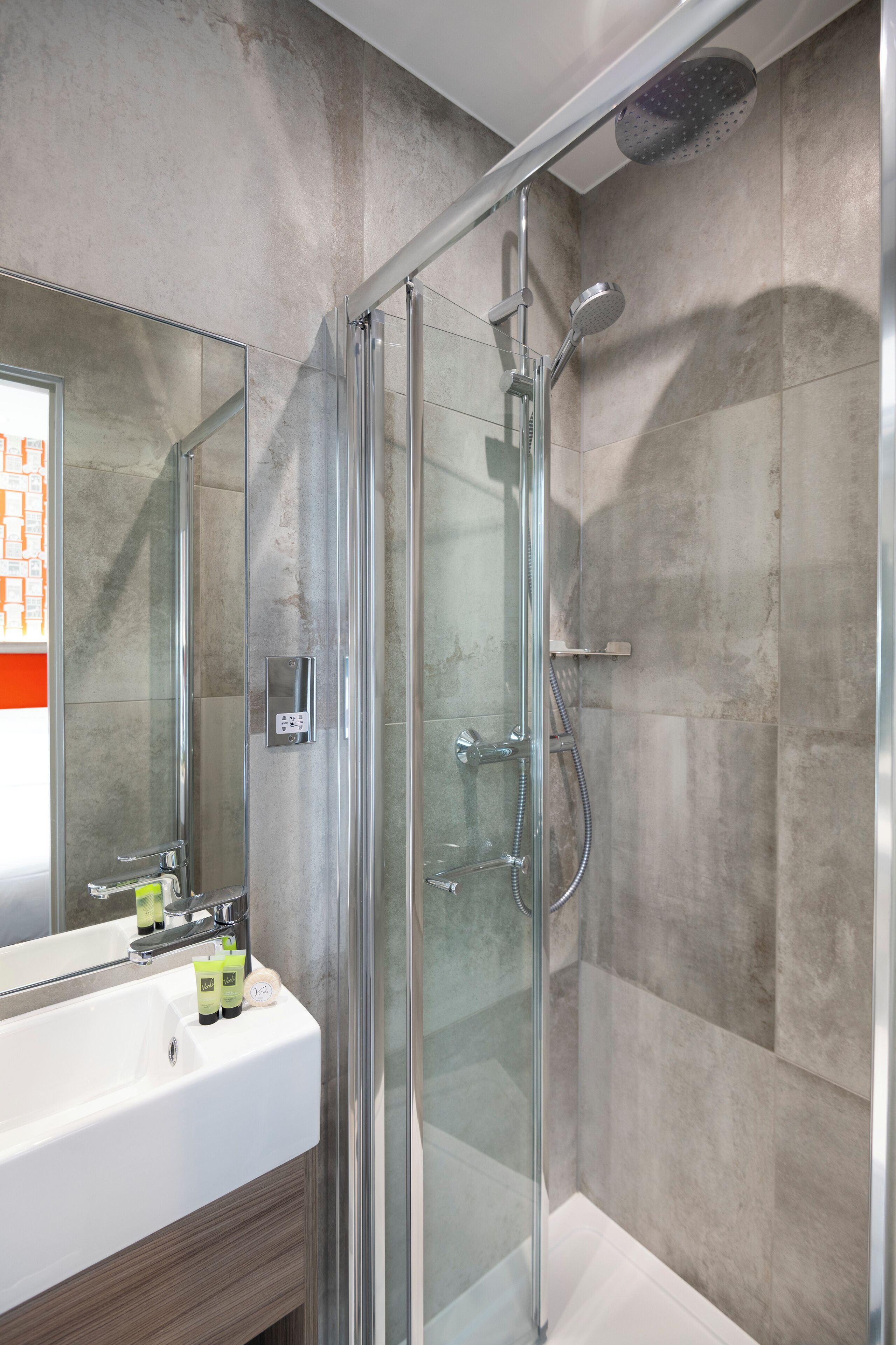 Un baño moderno con ducha de vidrio, un lavabo blanco con artículos de aseo y un espejo grande.