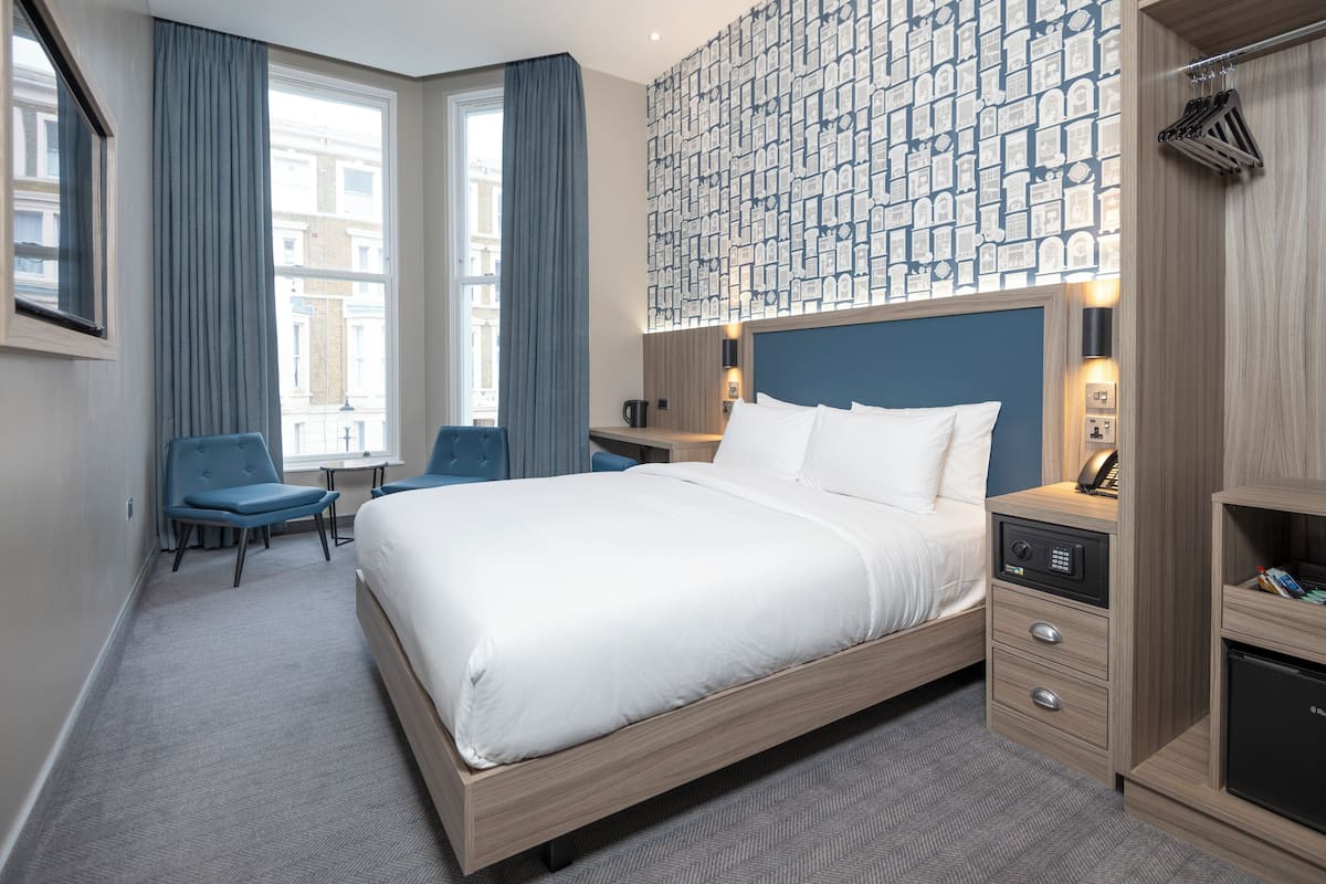 A hotel room with a large bed, a desk, two chairs, a wardrobe, and a patterned wall.