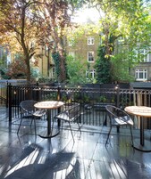 Een terras buiten met metalen stoelen en tafels, omringd door een zwart metalen hekwerk, en een uitzicht op een woongebouw op de achtergrond.