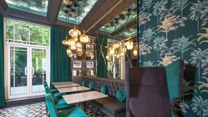 A restaurant with a high ceiling, green walls, and a patterned floor. There are multiple tables and benches, and a large window with a view of greenery outside. - Brit Hotels Earls Court (London)