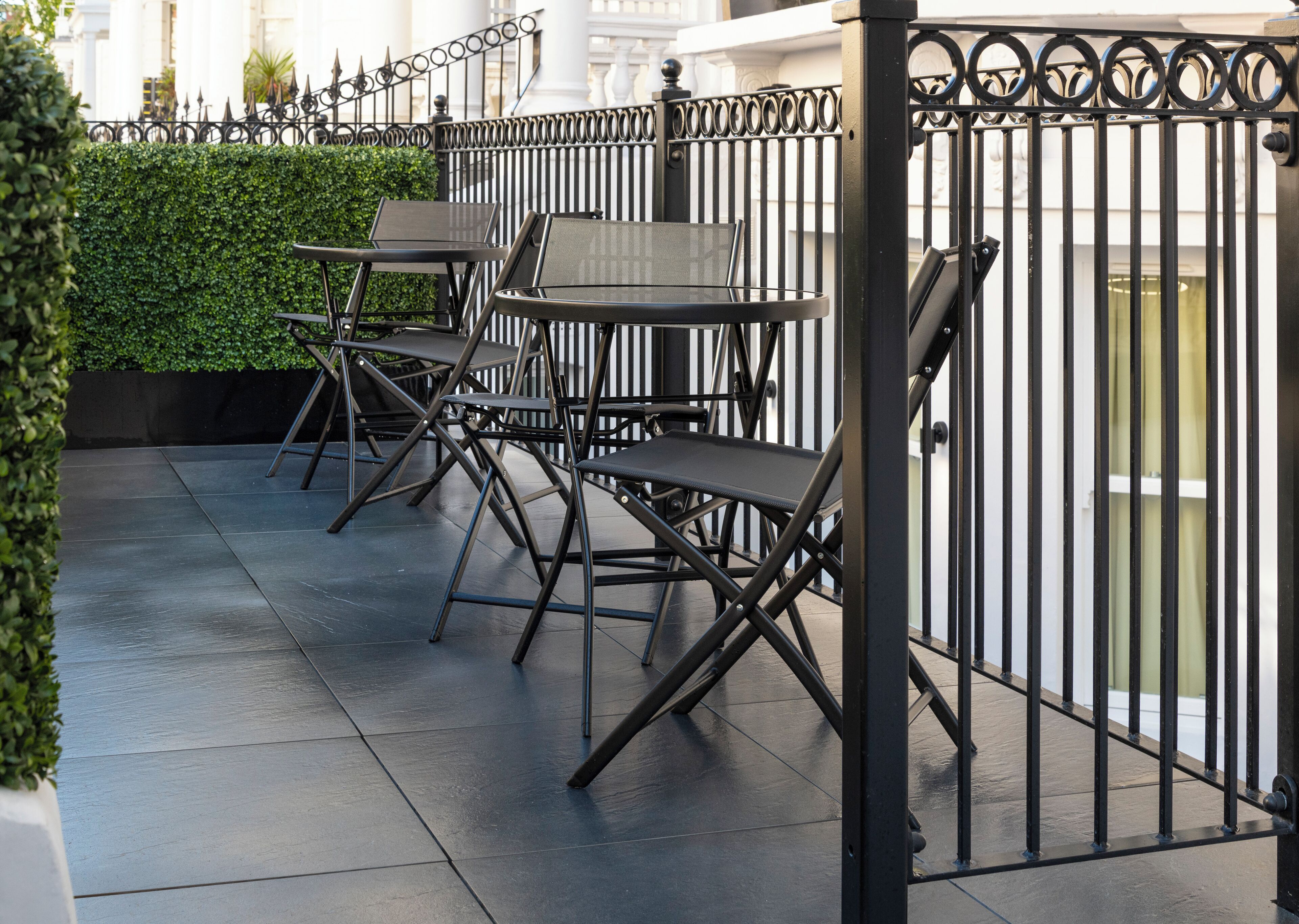 An outdoor seating area with tables and chairs, a metal fence, and a hedge.