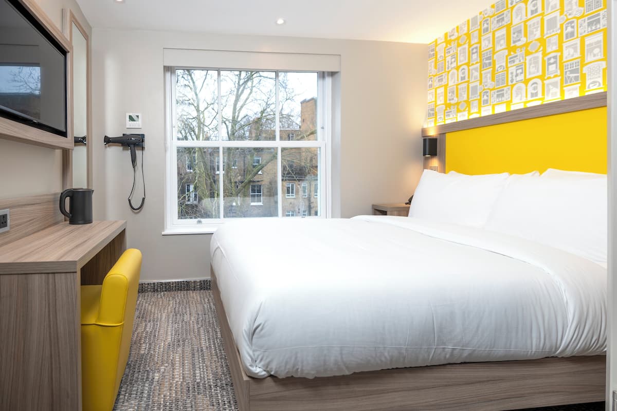 A hotel room with a bed, a desk, a chair, a coffee maker, a window, and a wall with a patterned design.