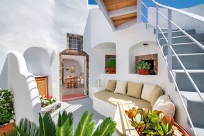 A modern white-walled room with a sofa, a staircase, and potted plants. - Petit Village (Fira)