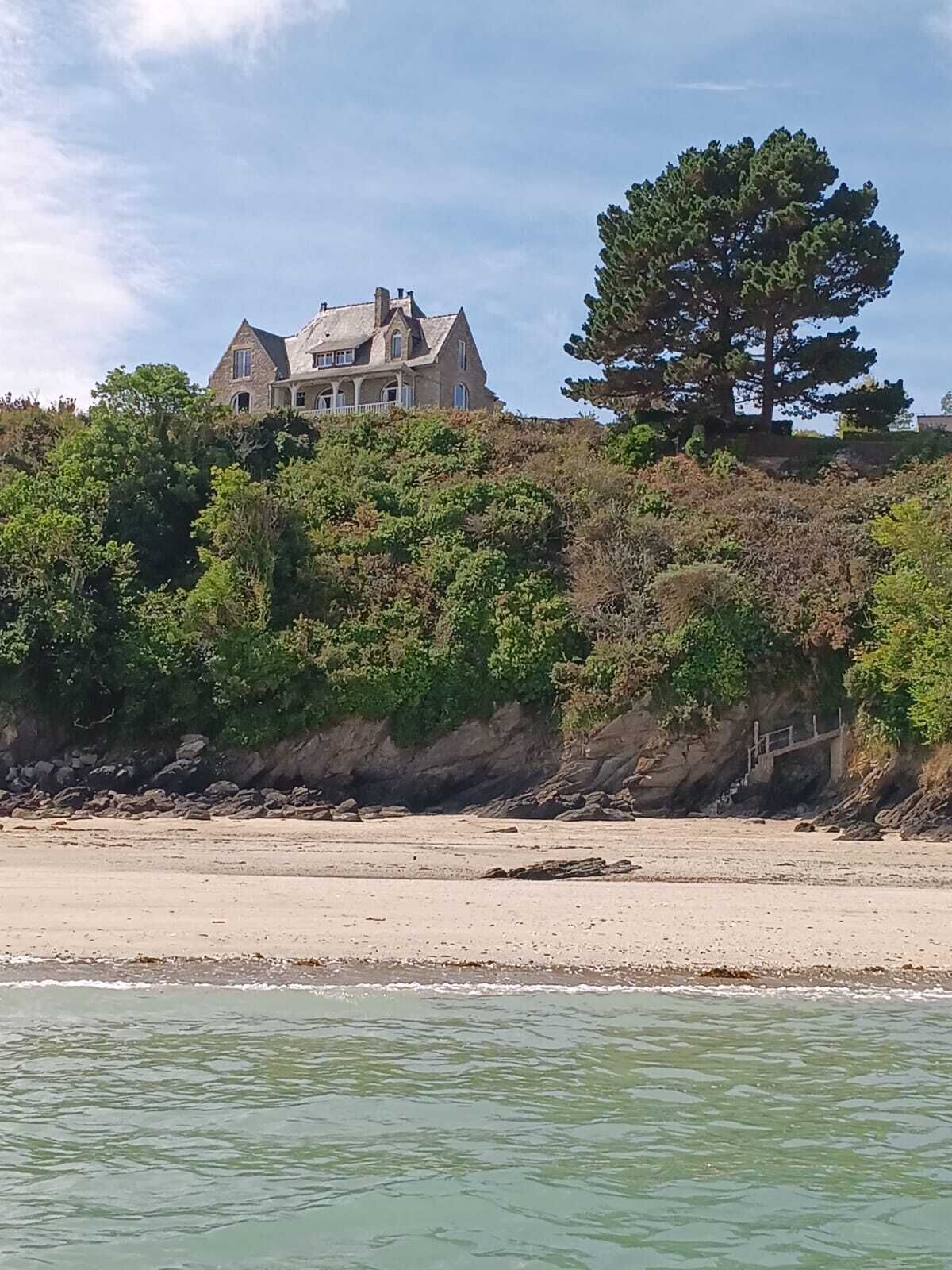 Une maison située sur la côte, avec vue sur la plage, entourée d’une végétation luxuriante et d’une falaise rocheuse.