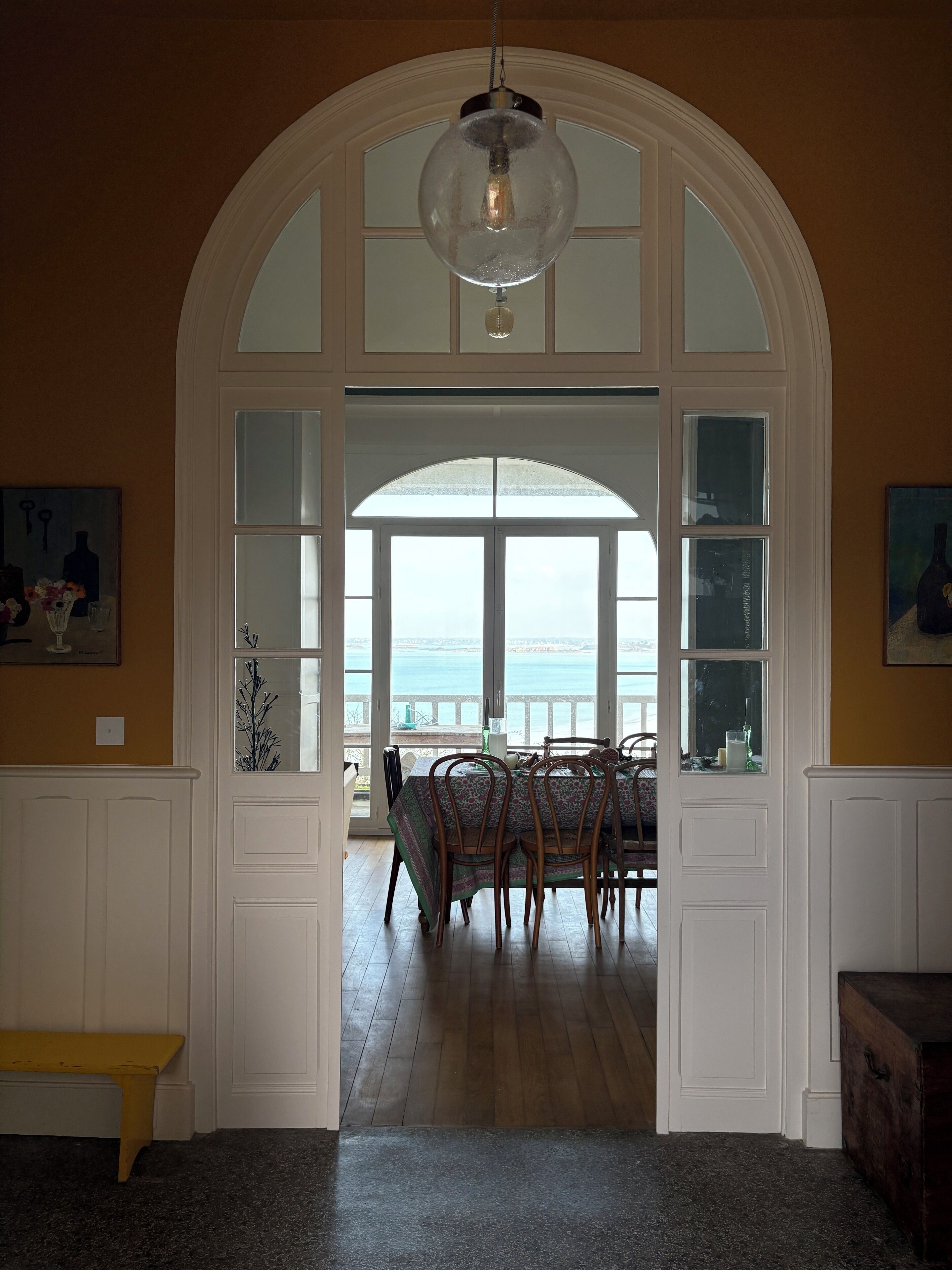 Un coin repas avec une table dressée pour quatre personnes, un banc et une vue sur la mer, visible à travers une grande fenêtre en arc de cercle.
