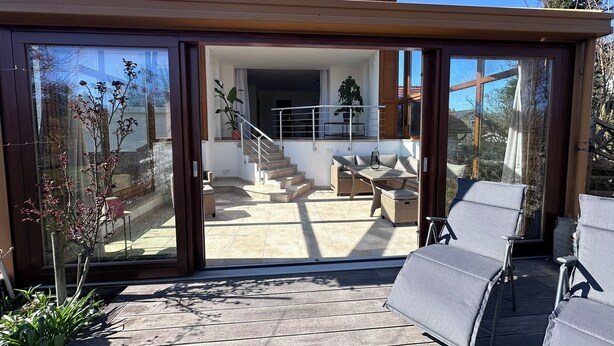 Een modern huis met een glazen deur die uitkomt op een terras met een bank en tafel.