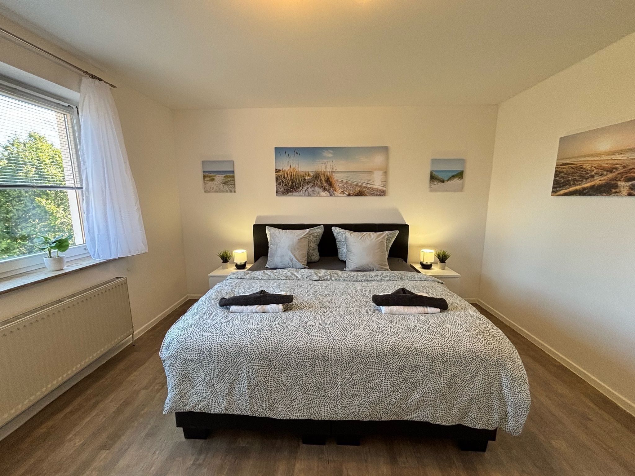 Een slaapkamer met een bed, twee nachtkastjes, een raam met gordijnen en drie foto's aan de muur.