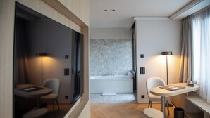 Une chambre d’hôtel moderne avec un plancher en bois, une petite table ronde avec une chaise, un canapé, un grand miroir, une fenêtre avec des rideaux et une salle de bain dotée d’une baignoire et d’un mur en marbre.