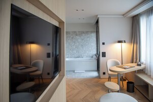 Habitación de hotel moderna con suelo de madera, una pequeña mesa redonda con una silla, un sofá, un espejo grande, una ventana con cortinas y un baño con bañera y pared de mármol.