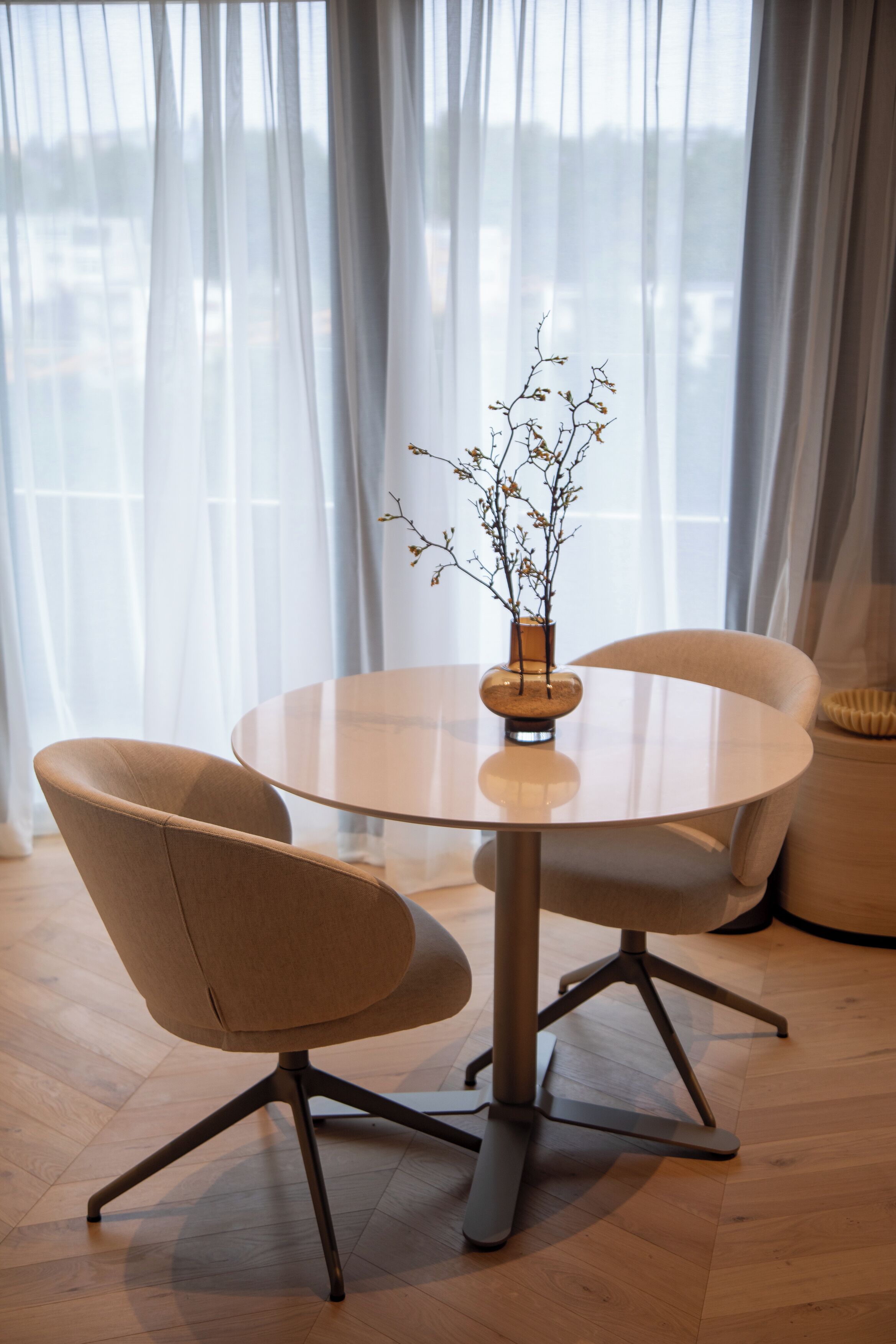 A round table with a vase of branches, two chairs, and a view of a city through sheer curtains.