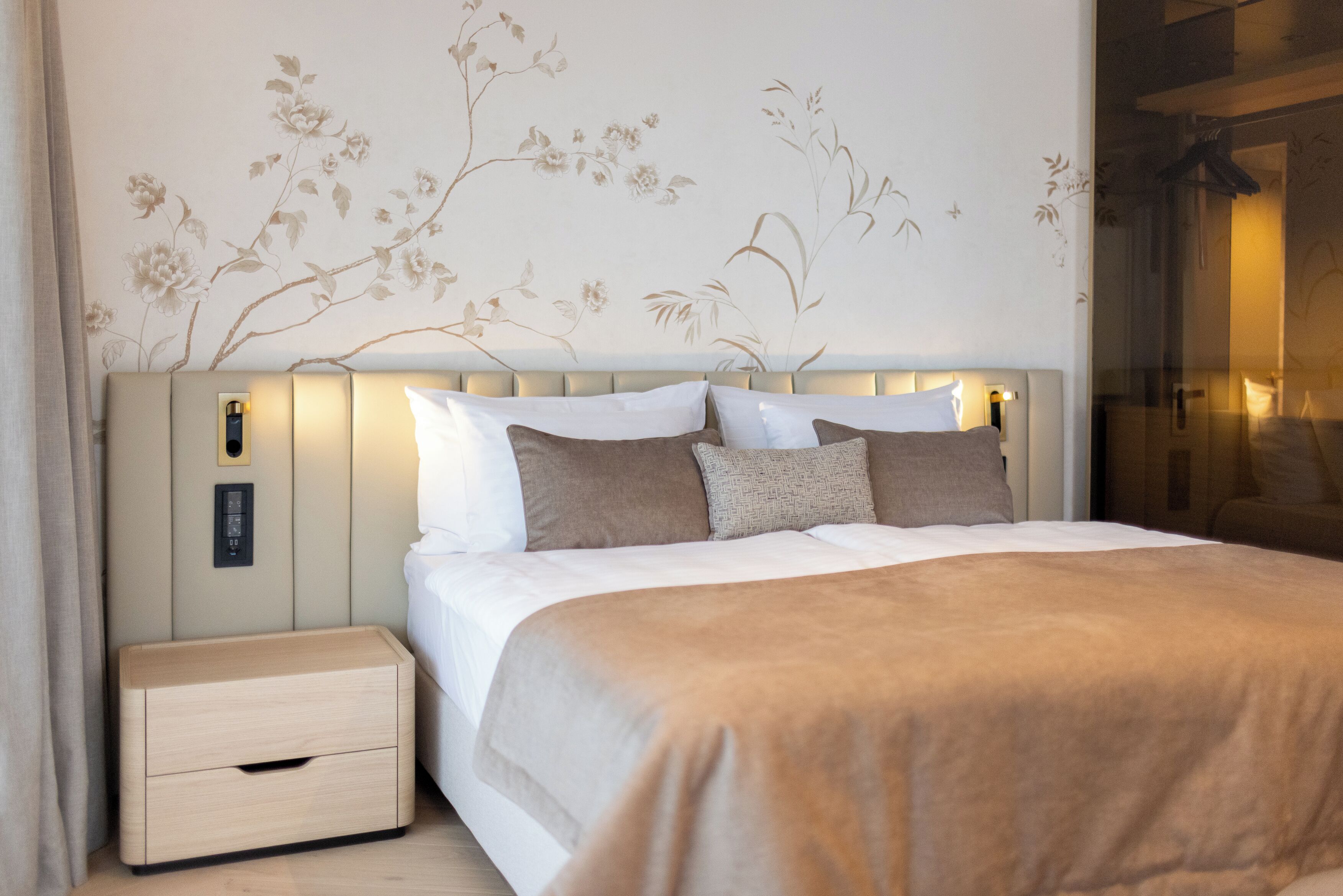 Una cama bien tendida con cabecera color beige, una mesita de noche y una pared decorada con motivos florales.
