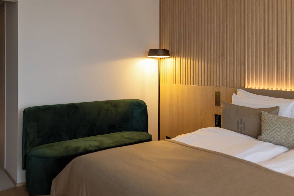Une chambre d’hôtel avec un lit, un banc recouvert d’un tissu vert, une table de chevet avec une lampe et une tête de lit en bois.