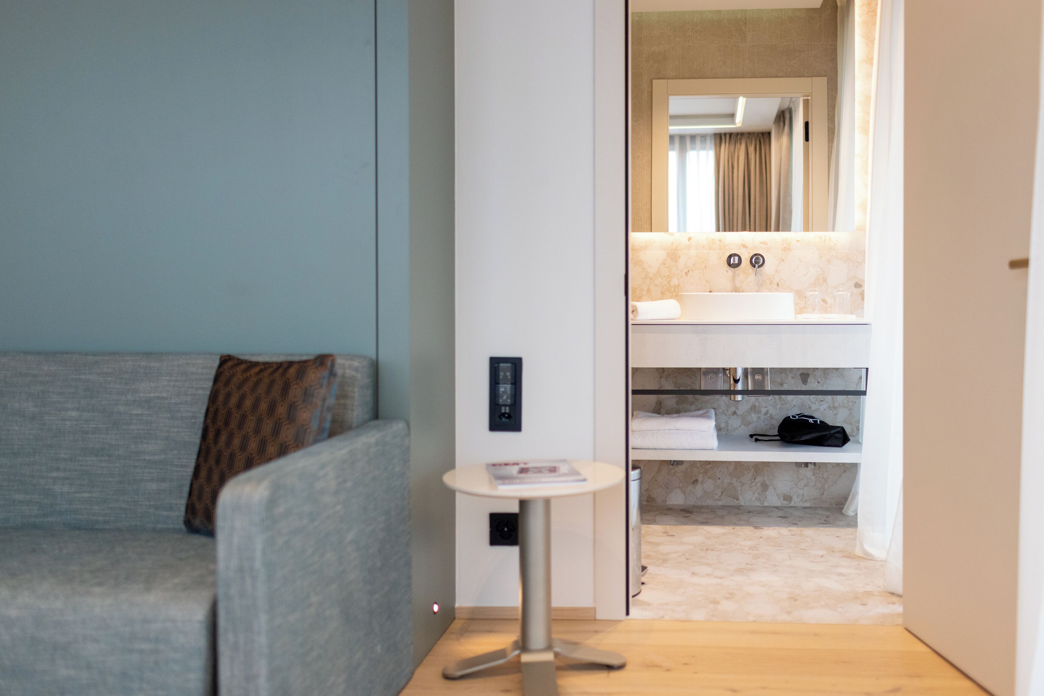 A modern hotel room with a grey sofa, a small round table, and a bathroom with a sink and mirror.