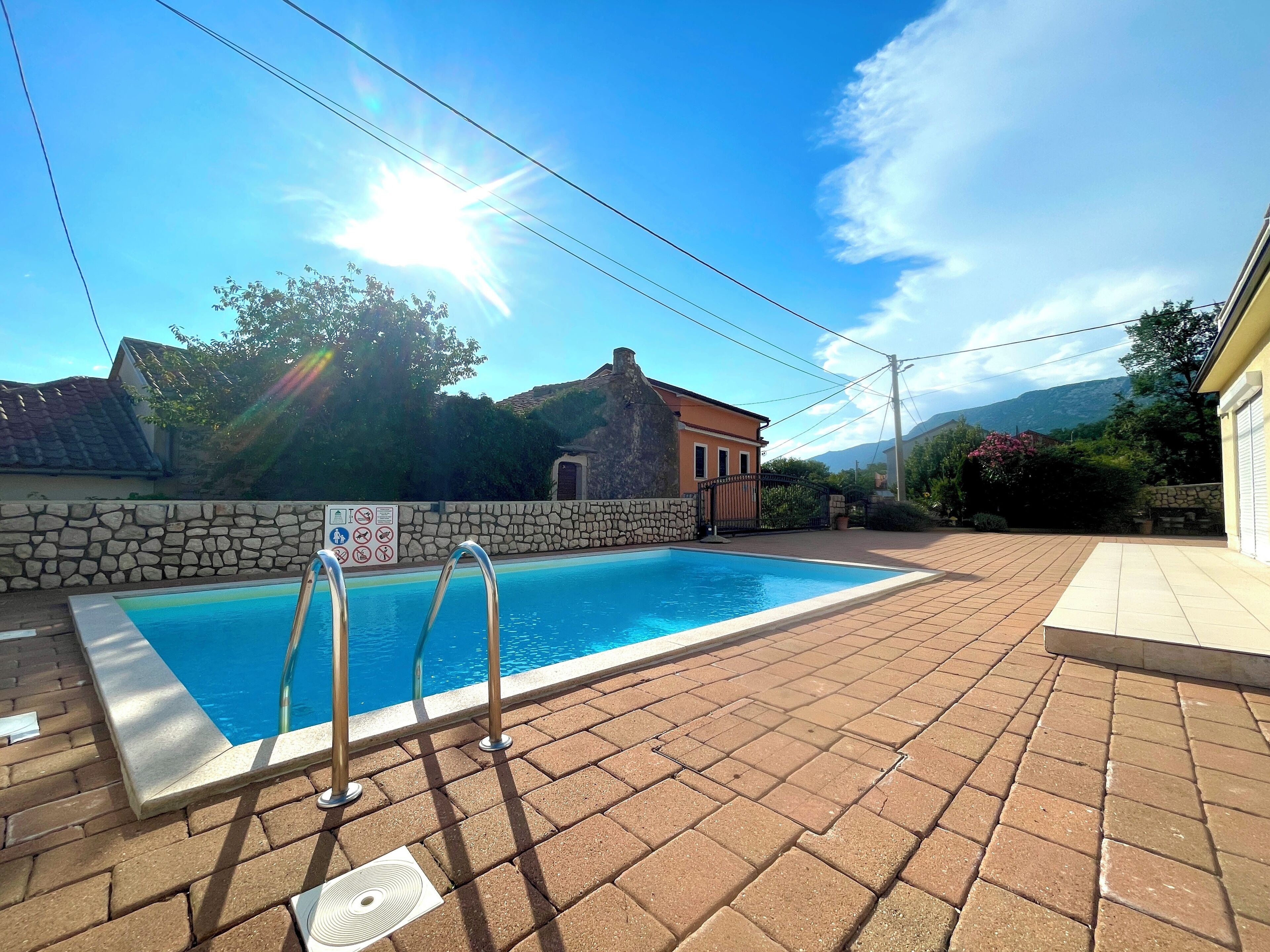 A swimming pool with metal ladders, surrounded by a stone wall and a paved area.